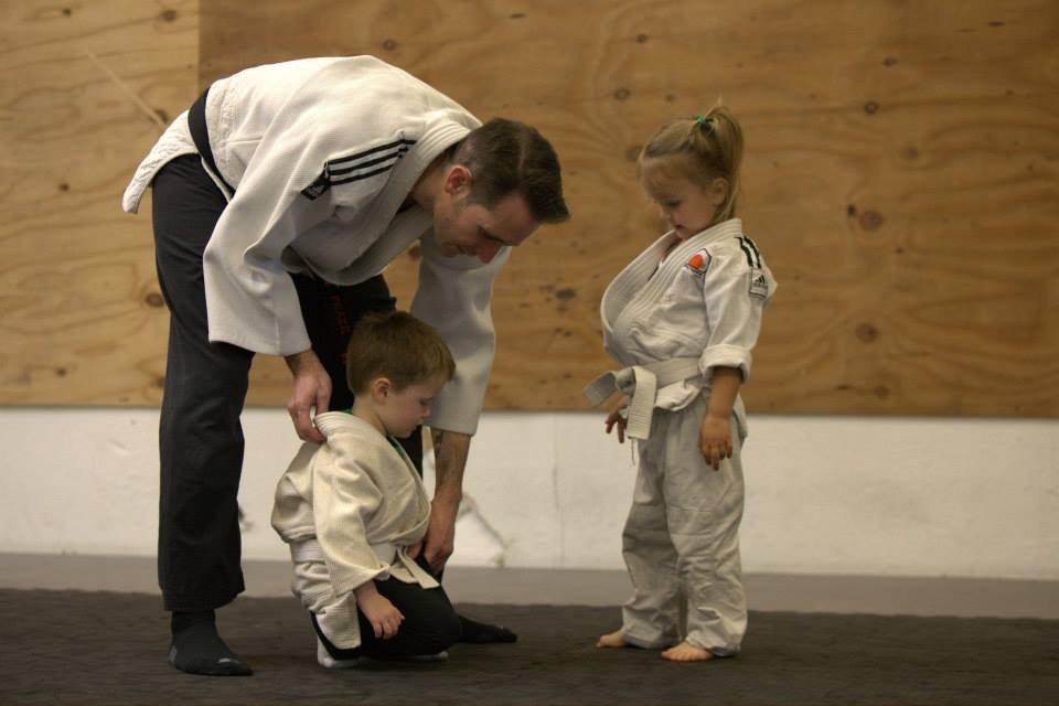 Coach Chris kneels to fix a small student's belt while another waits