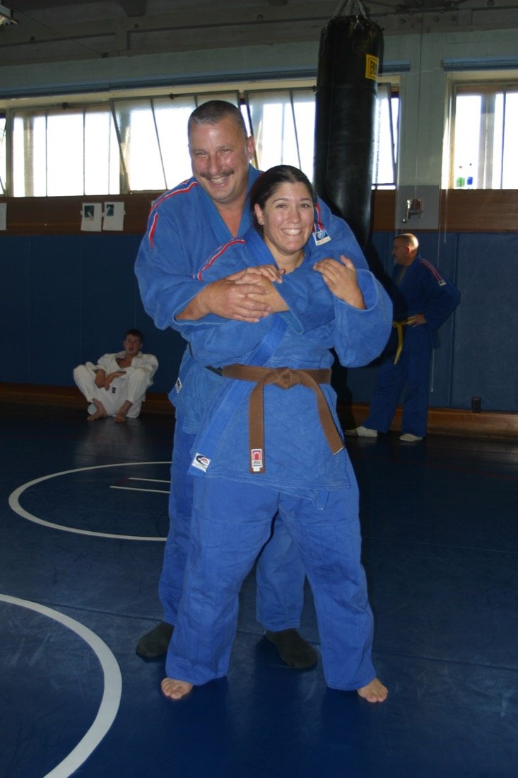 Coach Terry Kelly, smiling, with his arms around Coach Amy Carlisle, both in blue judogi on the mat at the SRJC gym