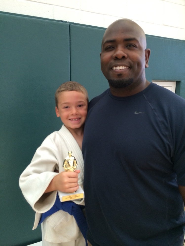 A young judoka holds a trophy next to Coach Dennis